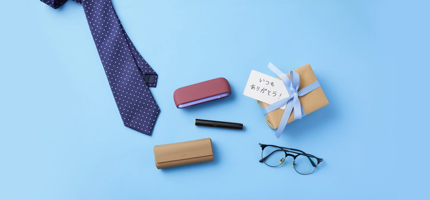 青地の背景に「いつもありがとう！」のメモがついた小包、ネクタイ、眼鏡、IOOS イルマ、IQOS イルマ プライムが置いてある様子