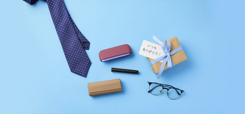 青地の背景に「いつもありがとう！」のメモがついた小包、ネクタイ、眼鏡、IOOS イルマ、IQOS イルマ プライムが置いてある様子